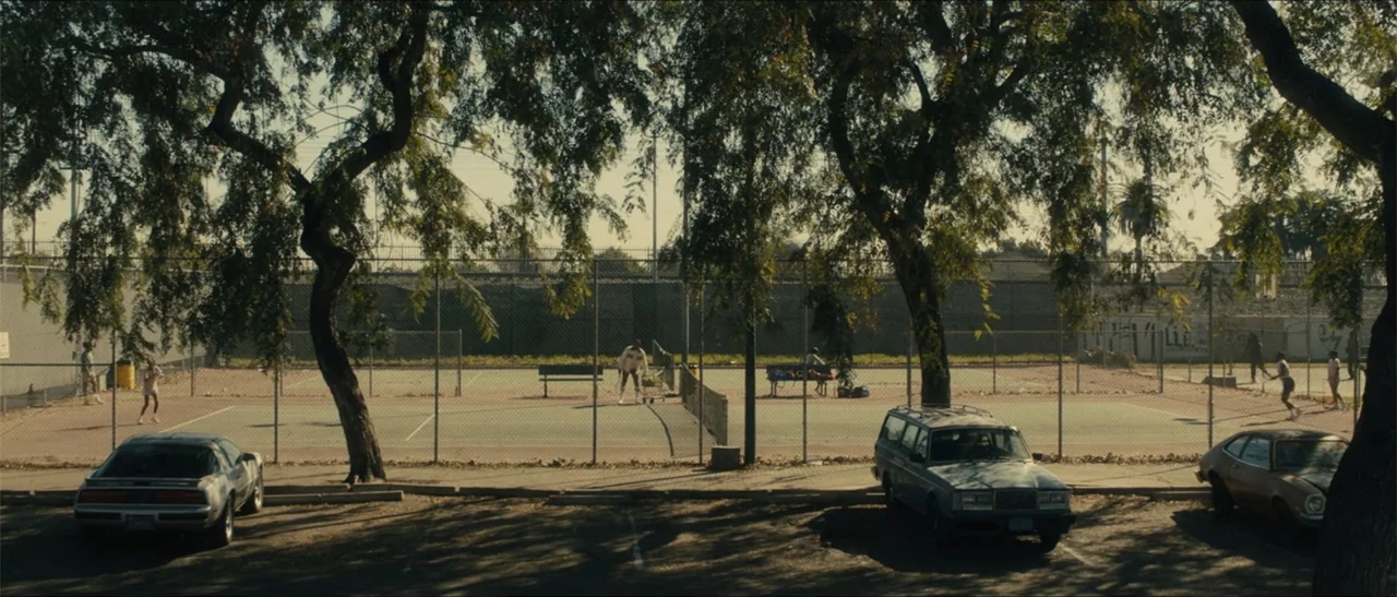 King Richard (2021) movie still: wide — An urban outdoor tennis court is enclosed by a chain link fence with trees. Several people play tennis in…