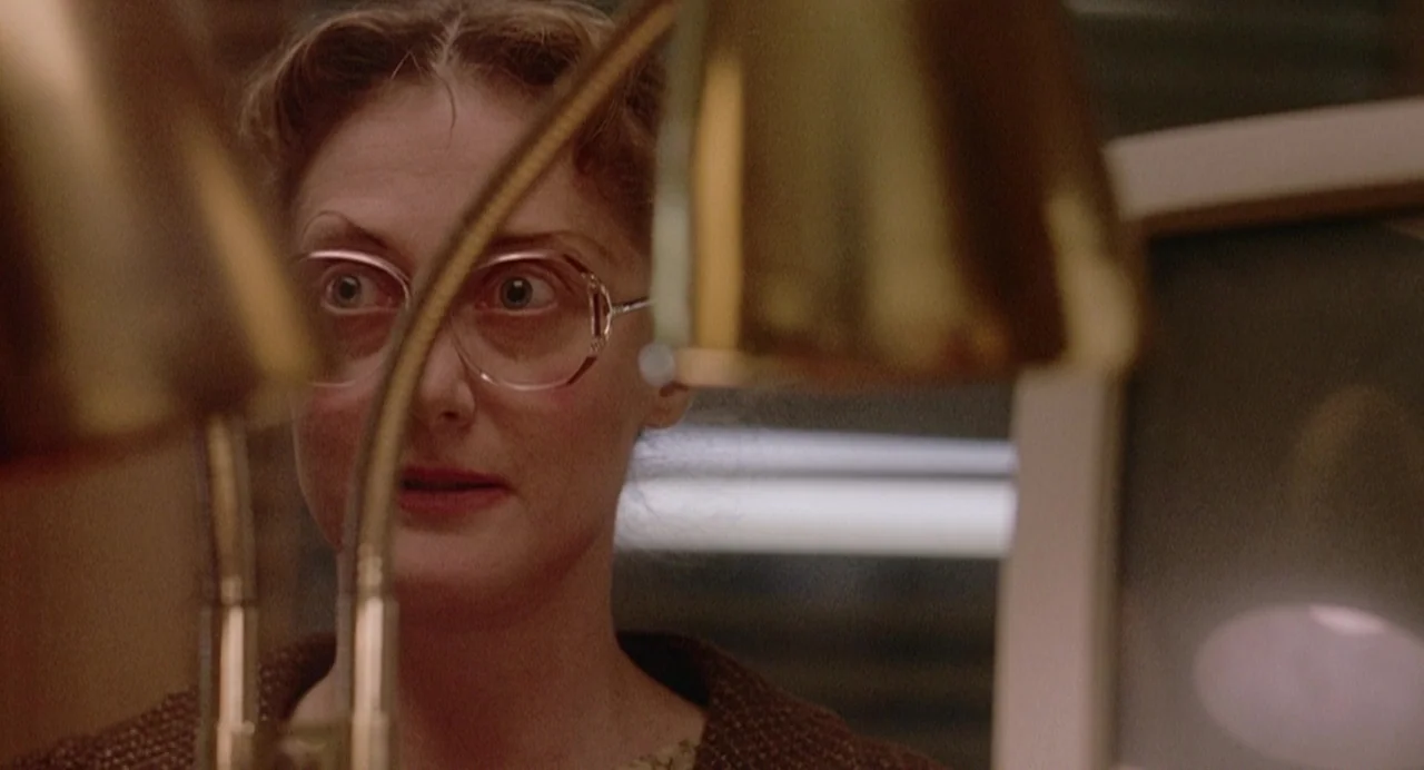 Office Killer (1997) movie still: closeup — An interior office space with metallic lamps positioned in the foreground. A woman looks forward through…