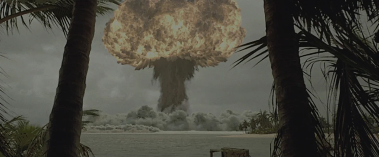 Godzilla (2014) movie still: wide — A view of an ocean coastline through palm trees shows a nuclear explosion rising. A towering, billowing mushroom…