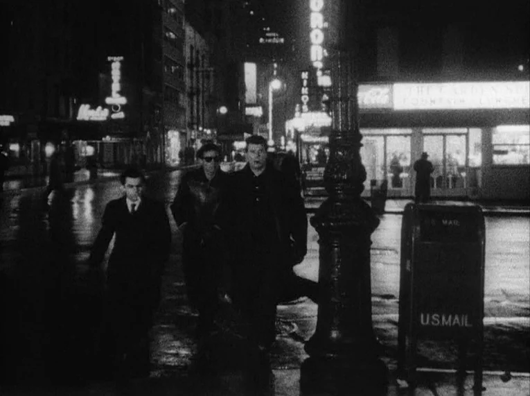 Shadows (1958) movie still: fullbody — A wet, dark city street at night illuminated by flickering neon store signs. Three men walk side-by-side past…