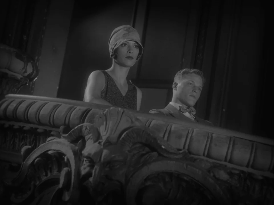 The Artist (2011) movie still: medium — A dark, ornate theater balcony with heavy stone carvings of ram heads in foreground. A young woman and man…