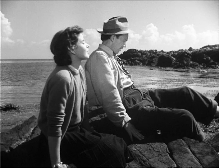 To Joy (1950) movie still: medium — Two people sit on dark jagged rocks by the ocean under a bright sky. They sit close together while facing toward…