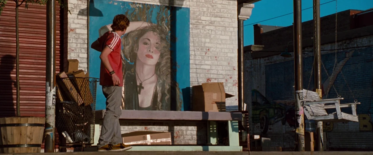 Empire Records (1995) movie still: A quiet, nostalgic moment where a young man in a red shirt pauses to look at a striking mural of a woman painted…