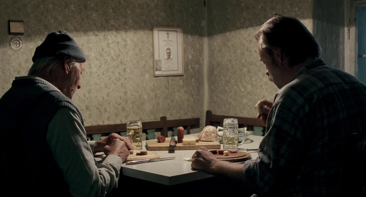 Revanche (2008) movie still: medium — A small dining area with patterned wallpaper and a framed portrait on the wall. Two men sit at a table…