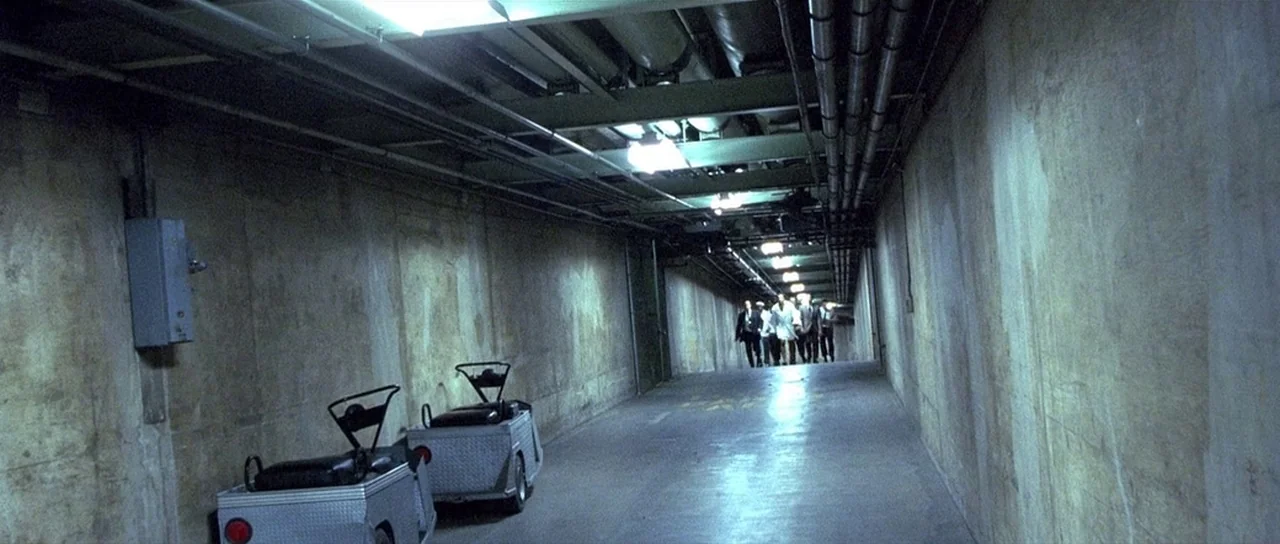 Ali (2001) movie still: wide — A long, industrial service tunnel features exposed concrete walls and ceiling piping. A group of individuals walks…