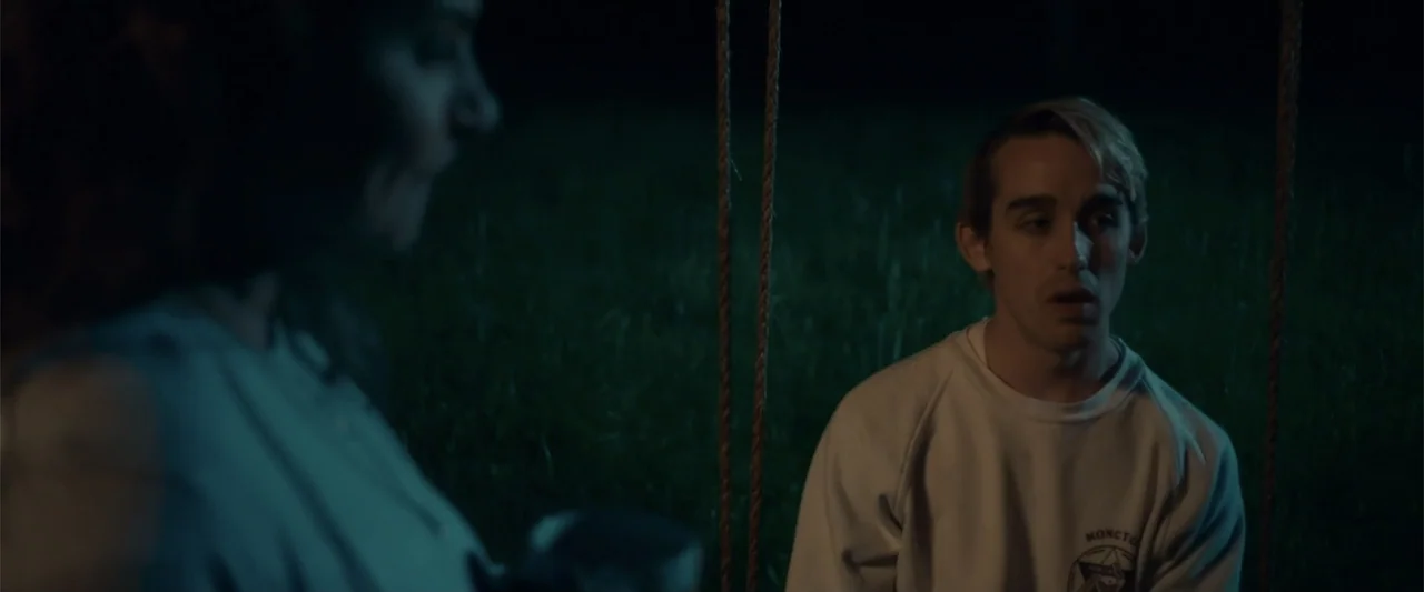 Jump, Darling (2022) movie still: medium — A night scene at a playground with a man sitting on a swing. The man looks toward someone off-camera in…