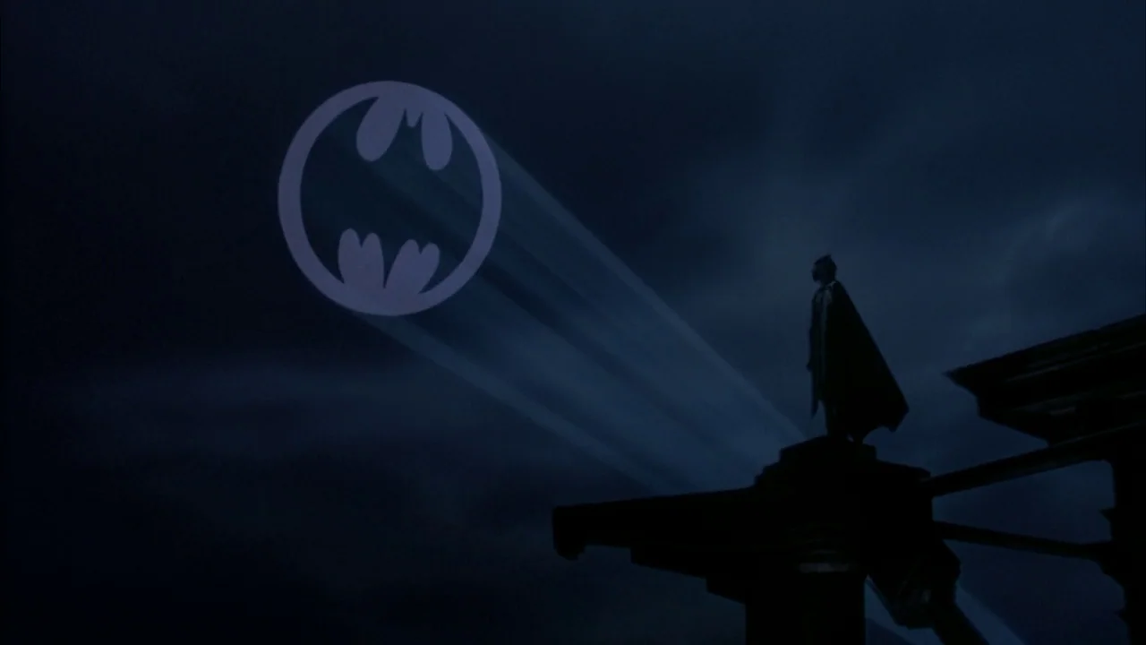 Batman (1989) movie still: wide — A dark night sky shows a large bat-shaped light beam projection against the clouds. A masked figure in a cape…