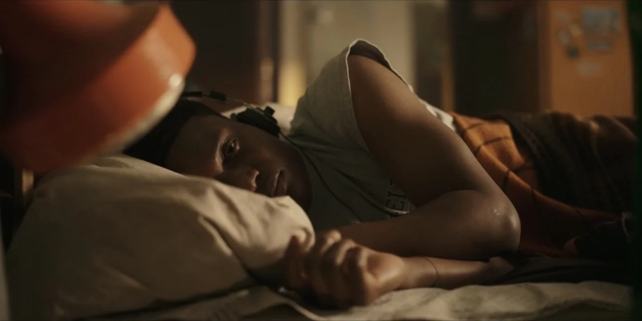 Autumn Beat (2022) movie still: medium — A dimly lit bedroom features a person lying in bed on a pillow. A young man with headphones rests his head…