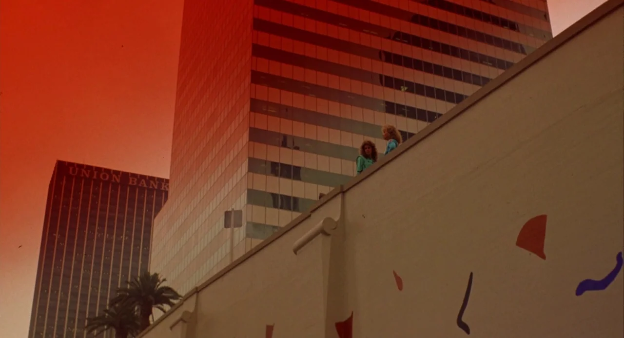 Night of the Comet movie still: wide — A low-angle view shows two women standing atop a ledge against tall modern skyscrapers. A large beige wall…