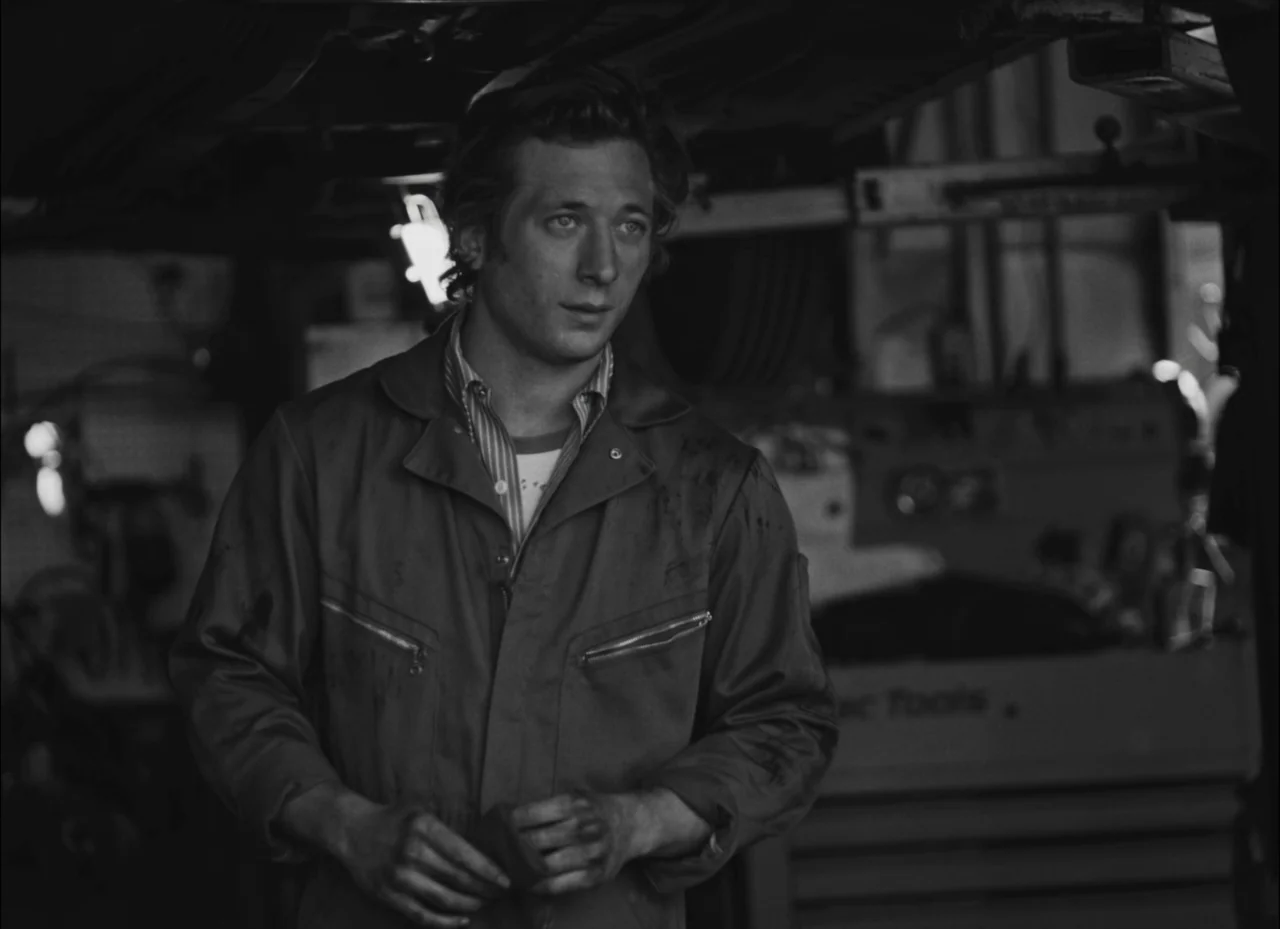 Fremont (2023) movie still: medium — An auto repair shop filled with tools and mechanical equipment in deep shadow. A man stands wearing a soiled…