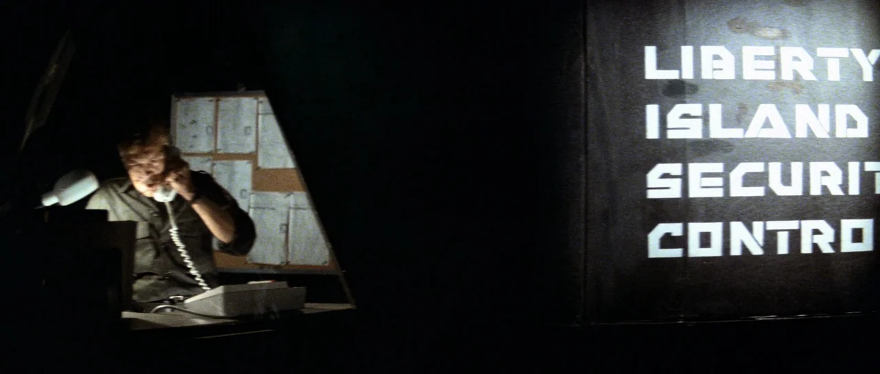 Escape From New York (1981) movie still: wide — A dimly lit room features a dark background wall with white block lettering. A man sits at a desk…