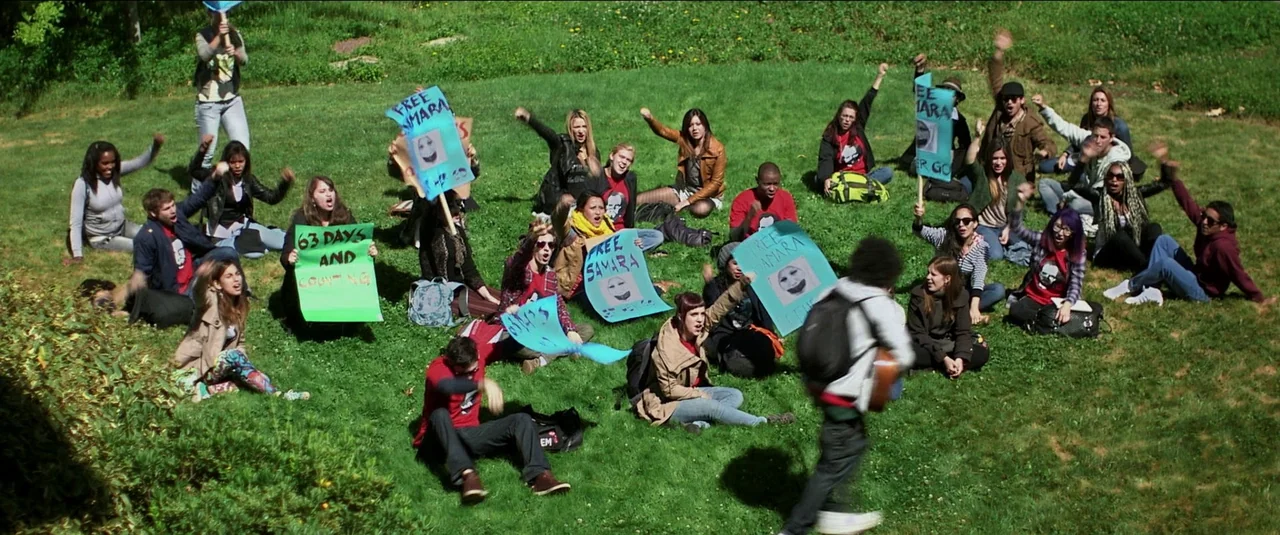 Green Inferno (2013) movie still: wide — A group of young people sit on a grassy hill holding signs with slogans. The individuals are raising their…