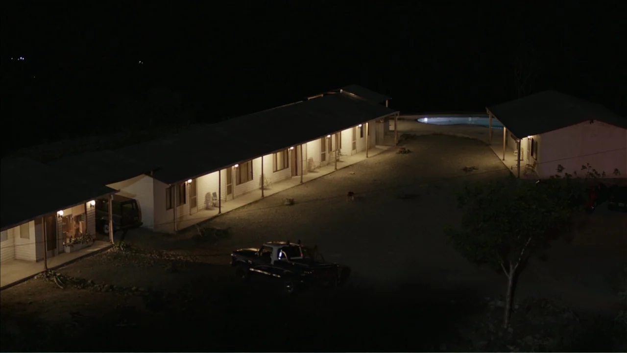 Boy (2010) movie still: establishing — A single-story motel building with a row of doors faces a dark dirt courtyard. A dark pickup truck is parked…