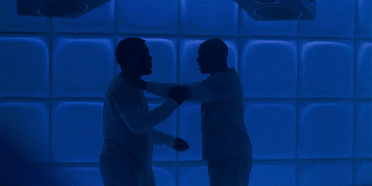 They Cloned Tyrone (2023) movie still: medium — A square-tiled padded room illuminated entirely by vibrant blue light. Two men in institutional…