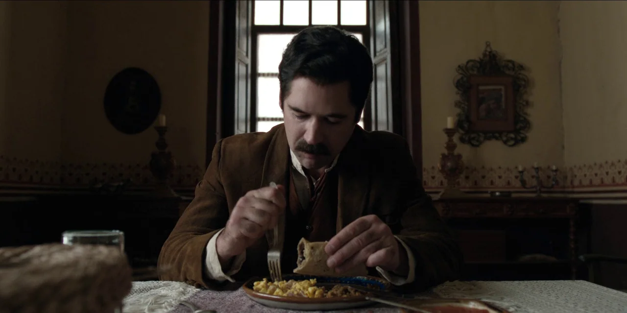 Pedro Páramo (2024) movie still: medium — A traditional dining room with antique furnishings and patterned wallpaper. A man in a brown jacket eats a…