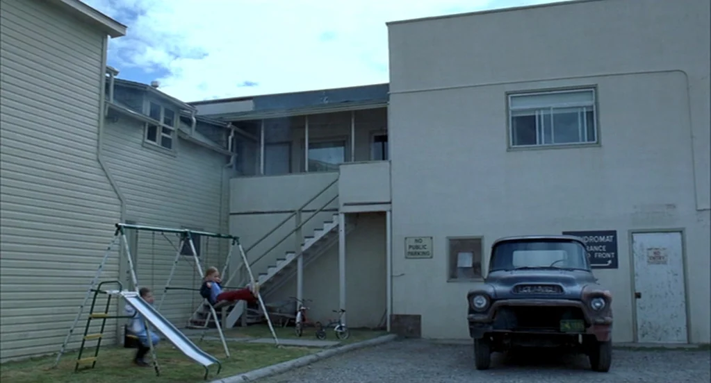 Brokeback Mountain (2005) movie still: wide — An outdoor courtyard sits between light-colored apartment buildings with an exterior staircase. Two…