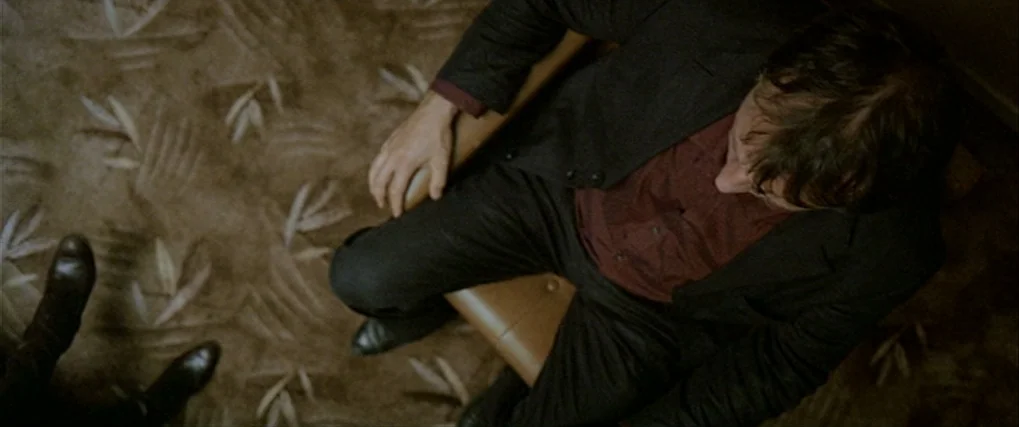 Next Door (2005) movie still: medium — A man sits in a chair atop a patterned brown carpet. He looks downwards while resting his hand on his knee.