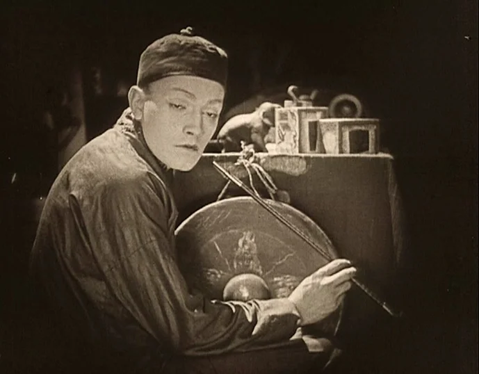 Broken Blossoms (1919) movie still: medium — An interior space features a table with small decorative items and a large gong. A man wearing a…