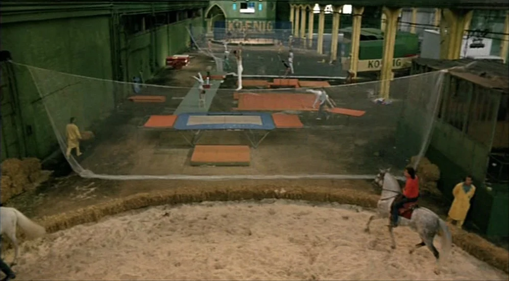 Roselyne and the Lions (1989) movie still: wide — A vast indoor training facility features green walls and a sandy equestrian ring. Several…