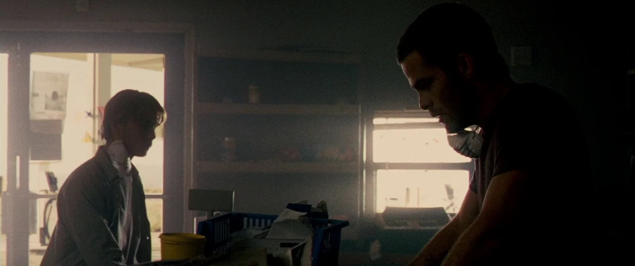 Carriers (2009) movie still: medium — A dark gas station interior is illuminated by bright daylight spilling through glass doors. Two men wearing…