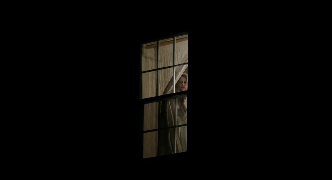 Antiviral (2012) movie still: wide — A dark night exterior features a single illuminated window centered in black negative space. A woman stands…