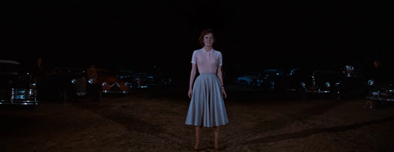 Rebel Without A Cause (1955) movie still: fullbody — A dark, gravel-covered outdoor area is surrounded by multiple parked 1950s automobiles. A young…