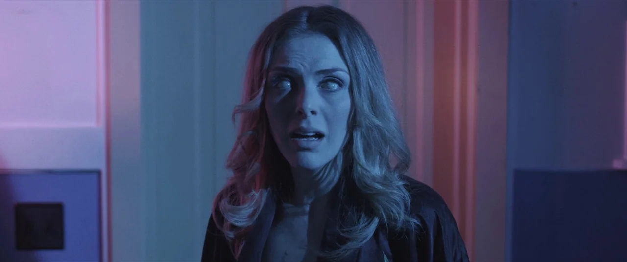 The Editor (2014) movie still: medium — A woman stands in a dimly lit hallway illuminated by magenta and blue light. She stares forward with milky…
