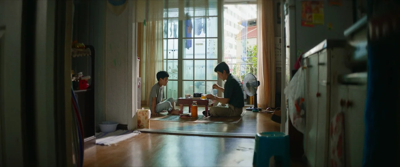 The Divine Fury (2019) movie still: wide — A modest apartment interior with a small dining table positioned before a large window. A man and a boy…