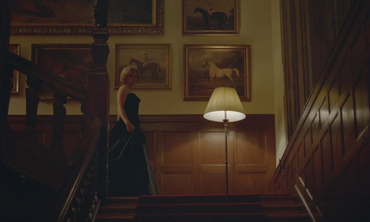 Spencer (2021) movie still: fullbody — A wooden staircase with a red carpet is adorned with framed oil paintings. A woman in a black gown walks down…