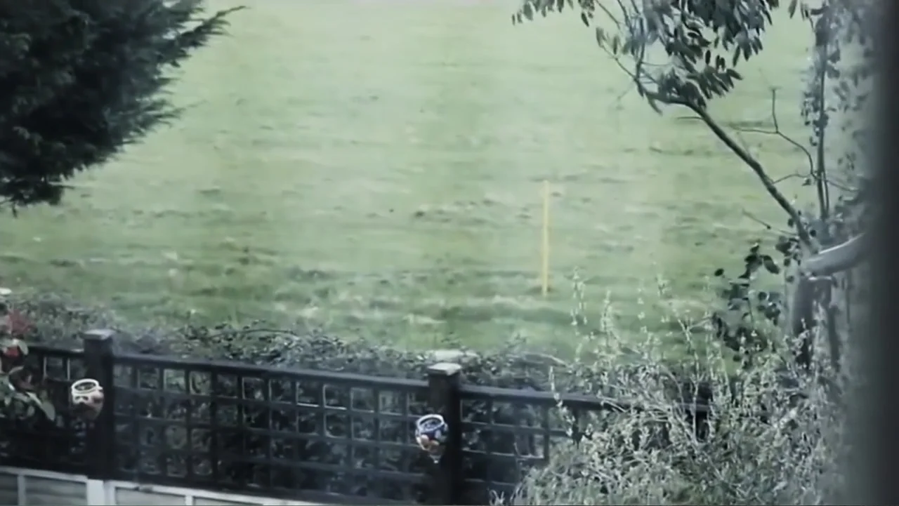 Beware the Slenderman (2016) movie still: establishing — A residential backyard overlooks a wide green field under an overcast sky. A black wooden…