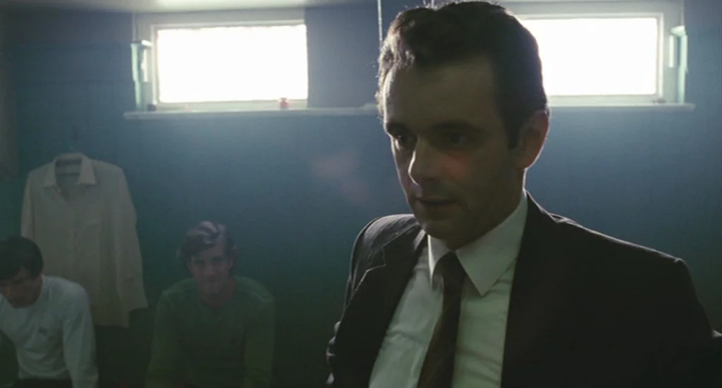 The Damned United (2009) movie still: medium — A dimly lit room with dark green walls and high, rectangular windows. A man in a suit stands in the…