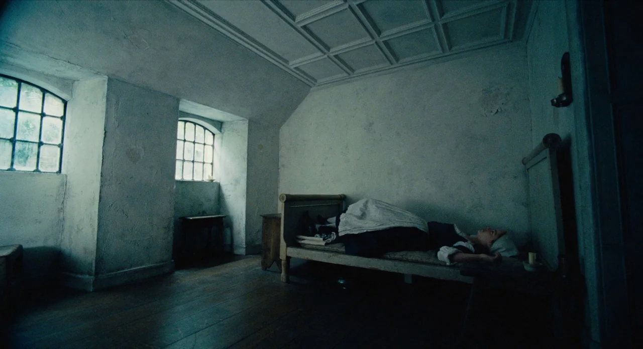 The Favourite (2018) movie still: wide — A austere, stone-walled room features two small windows with iron bars and low ceilings. A woman rests on a…