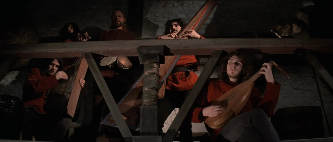 Macbeth (1971) movie still: medium — A group of musicians performs on a wooden balcony inside a dark stone room. The men wear matching red tunics…