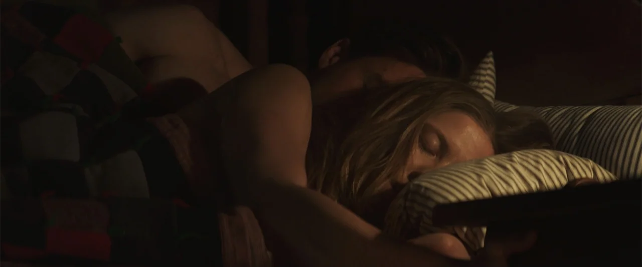 The Wind (2005) movie still: closeup — A dimly lit bedroom features a bed with a patterned quilt and striped pillows. A man and woman rest together…