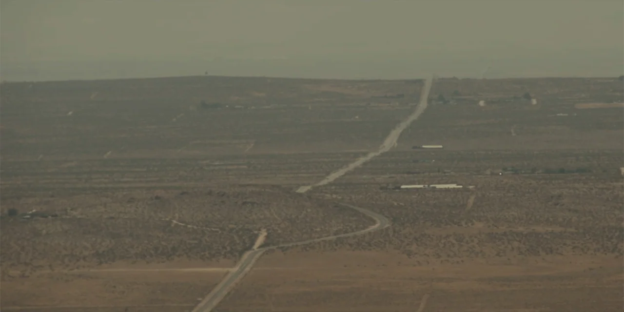 Fall (2022) movie still: establishing — A vast, arid desert landscape features a single road curving through sparse scrub vegetation. The highway…