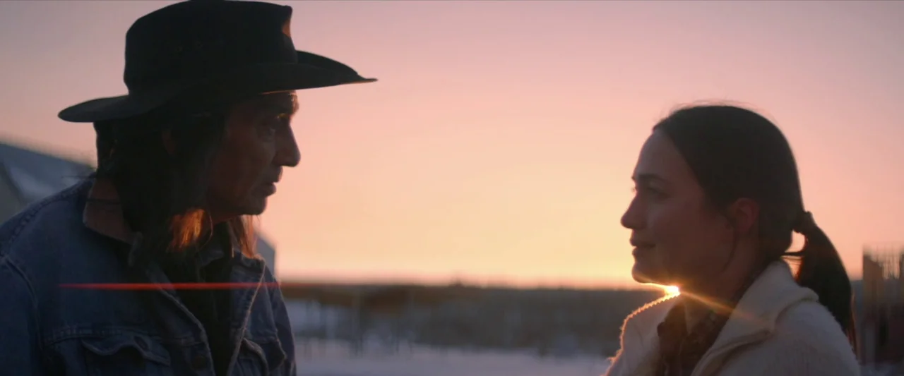 The Unknown Country (2022) movie still: medium — An open field covered in light snow during sunset with a soft, warm glow. An older man wearing a…