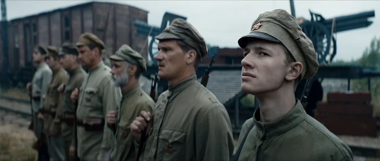 The Rifleman (2019) movie still: medium — A group of soldiers in 1940s-era uniforms stands on a gravel rail track. Several men in olive tunics are…