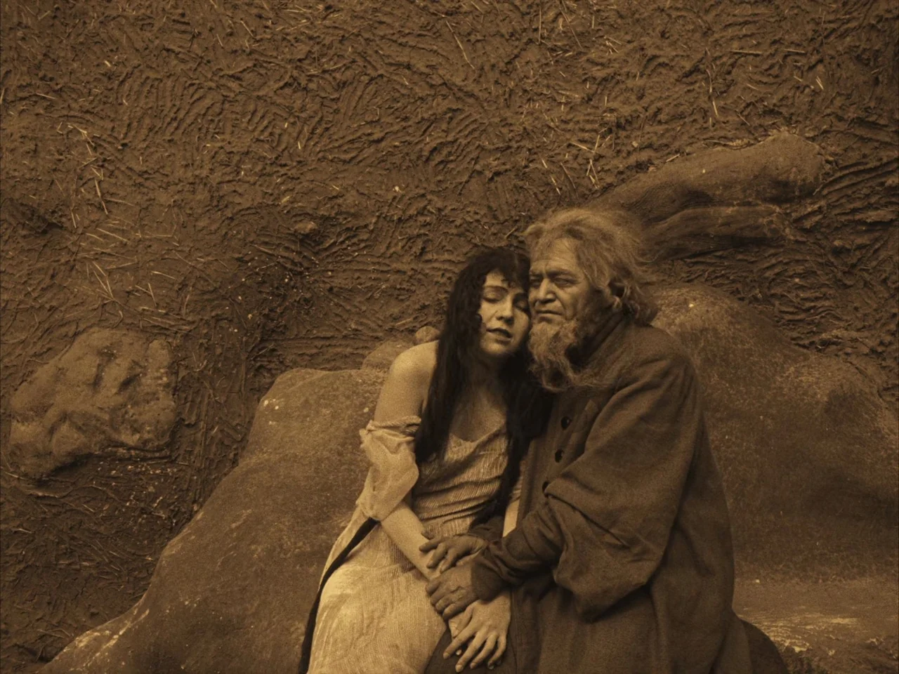 The Golem (1920) movie still: medium — Two individuals sit together against a rough mud-plastered or rock wall surface. The woman rests her head on…