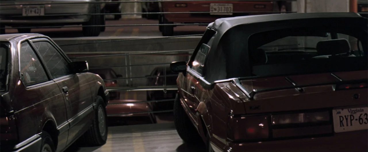 The Pelican Brief (1993) movie still: medium — A multi-level concrete parking garage contains several stationary automobiles at night. Two cars in…