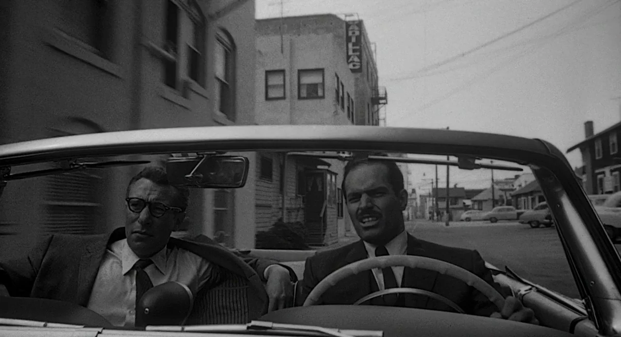 Touch of Evil (1958) movie still: medium — Two men sit in a convertible car driving through a quiet mid-century American suburb. The street is lined…