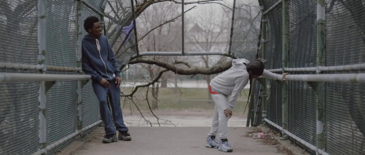 The Fits (2016) movie still: fullbody — An enclosed metal pedestrian walkway features chain-link fencing along both sides. Two young people stand…