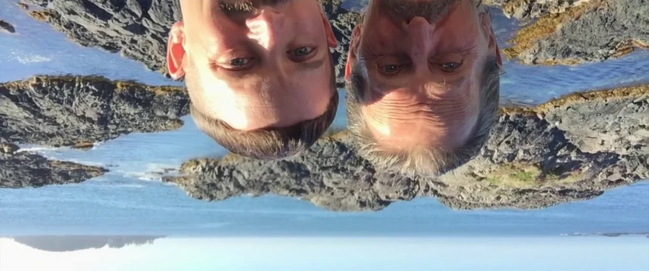 Come To Daddy (2019) movie still: closeup — An upside-down shot of two men in front of a rocky ocean shoreline. They are positioned close to the…