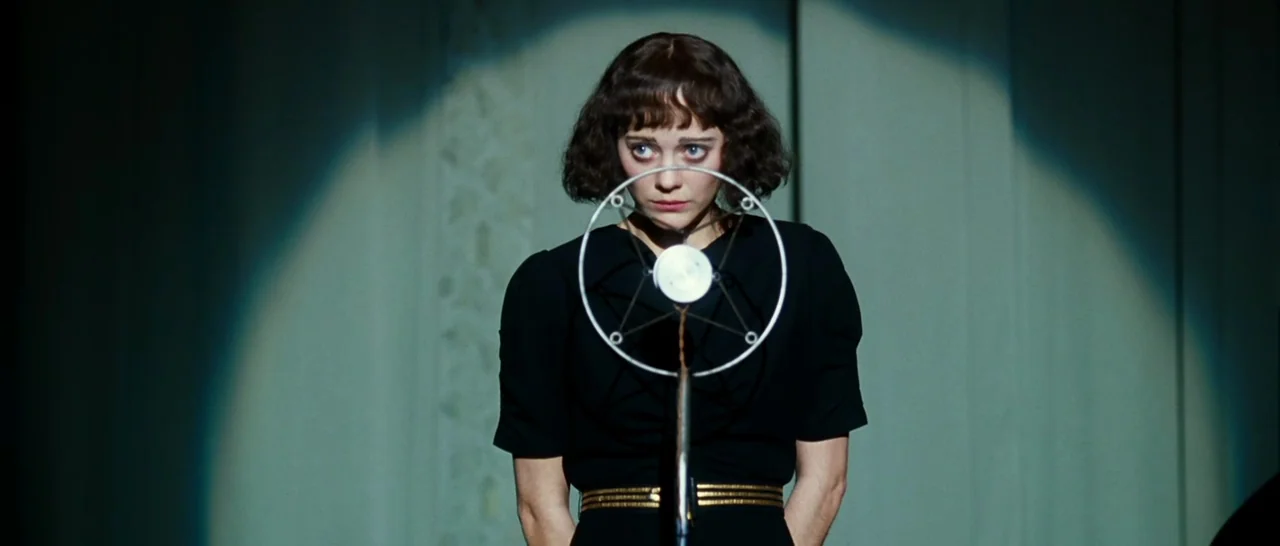 La Vie En Rose (2007) movie still: medium — A woman stands in front of a circular microphone on a plain studio set. She holds her hands behind her…