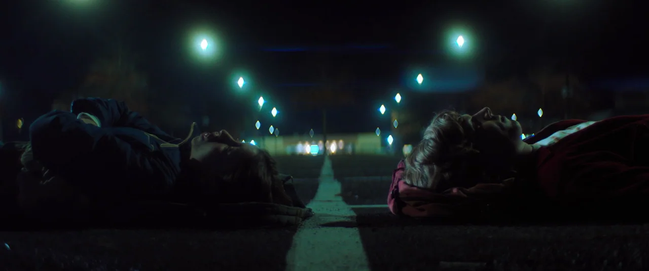 Linoleum (2022) movie still: wide — A dark asphalt parking lot at night with a painted white lane marker visible. Two teenagers lie on the ground on…