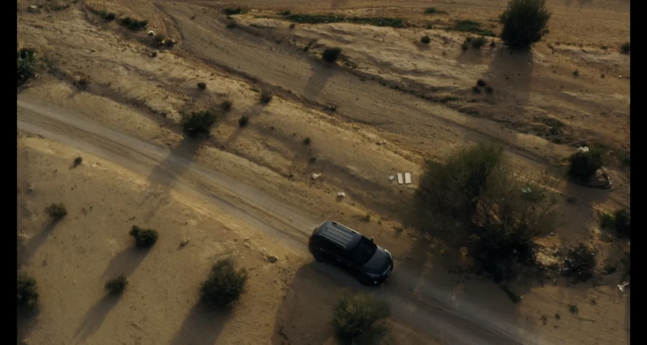 Saudi Women's Driving School (2019) movie still: establishing — A dark vehicle moves along a dusty track through a vast arid desert landscape. Low…