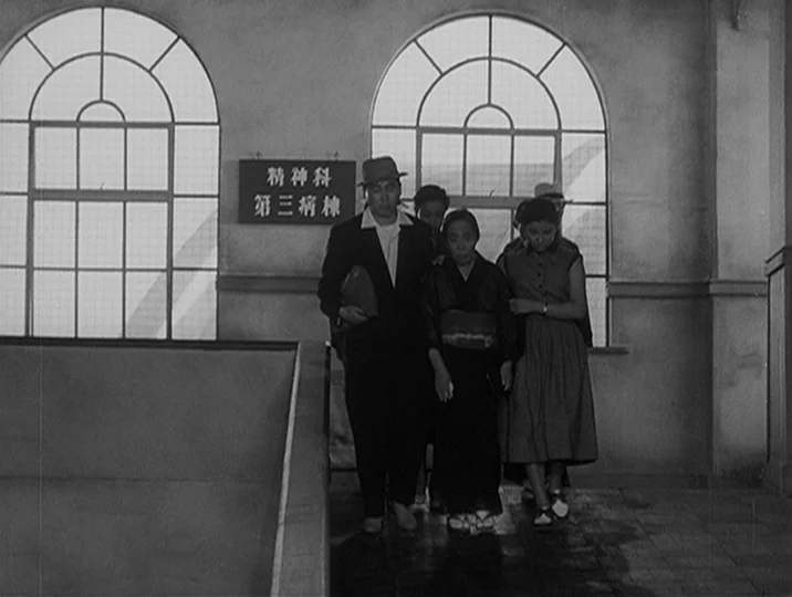 I Live In Fear (1955) movie still: fullbody — An institutional hallway features two large arched windows with gridded glass panes. A group of three…