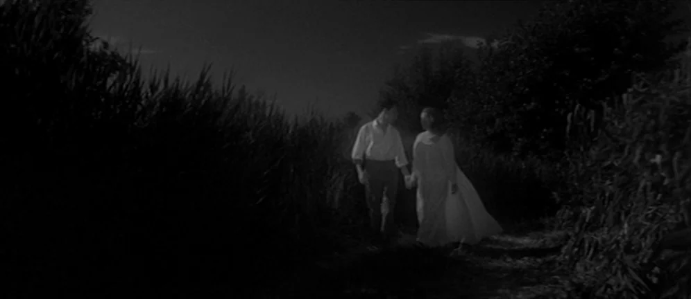Les Amants (1958) movie still: fullbody — A man and woman walk through a dark, overgrown field at night. They hold hands as they move along a narrow…