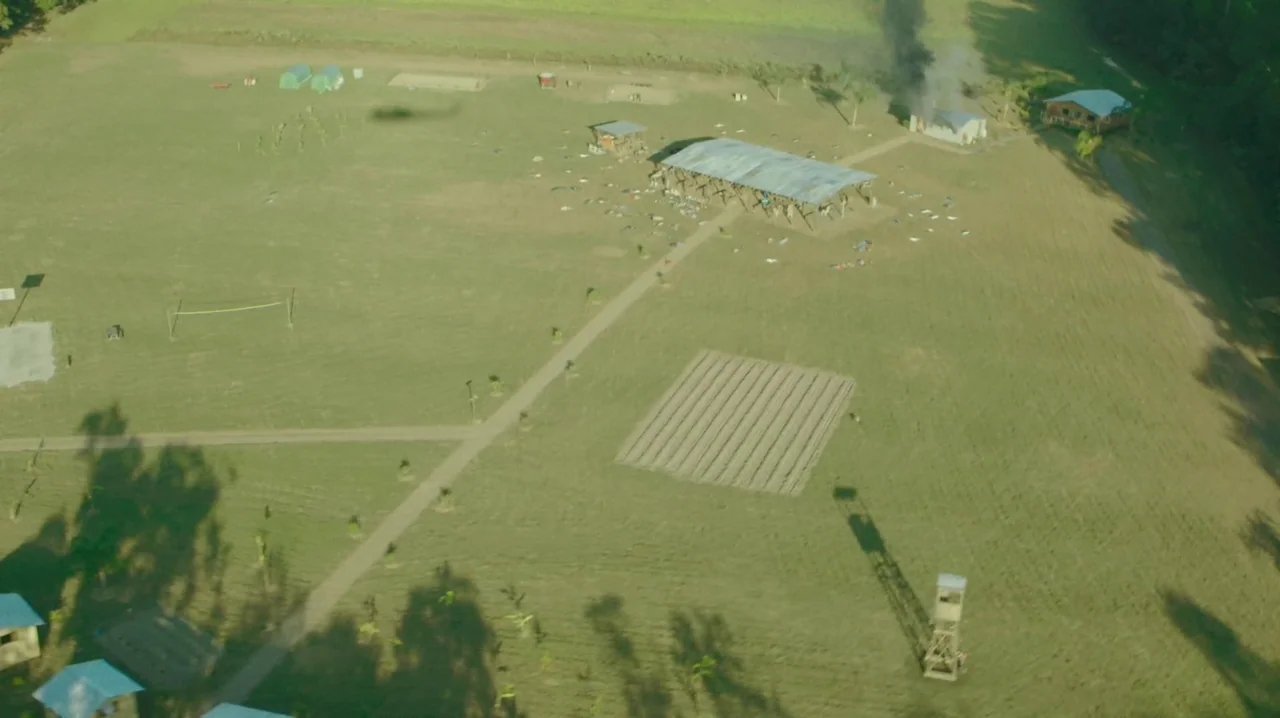 The Sacrament (2013) movie still: establishing — A rural compound featuring a large central pavilion and a small burning building. A dirt path…