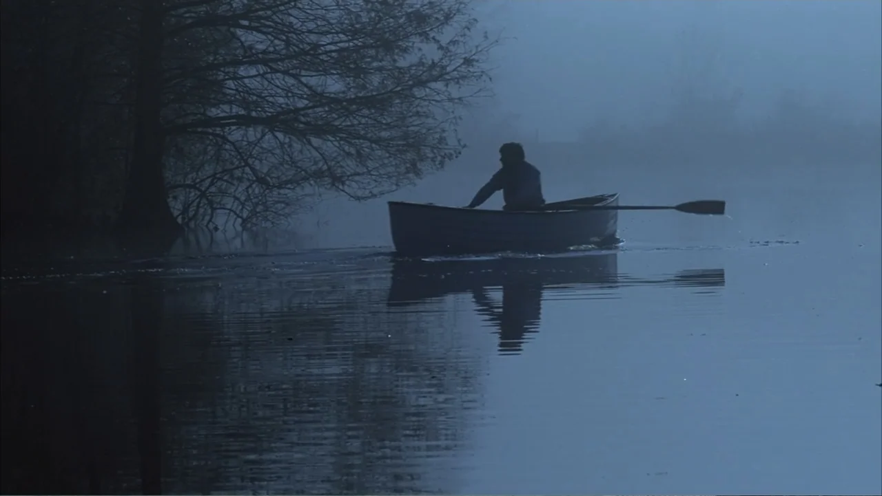 The Notebook (2004) movie still: wide — A calm lake is surrounded by dark, silhouettes of trees under a dim night sky. A person sits in a rowboat…