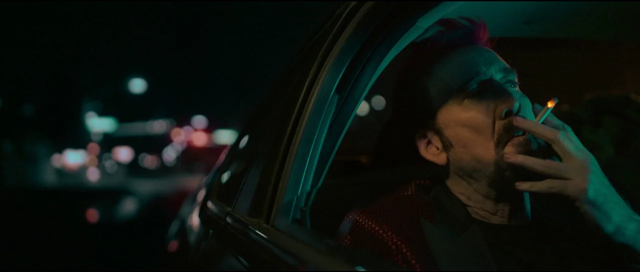 Sympathy For The Devil (2023) movie still: closeup — A man sits inside a dark car at night under neon street lights. He holds a burning cigarette in…